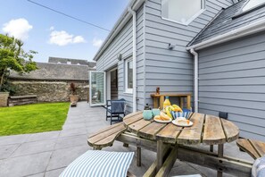 A patio with a wooden table, chairs, and a bench. There is a stone wall and a grassy area in the background.