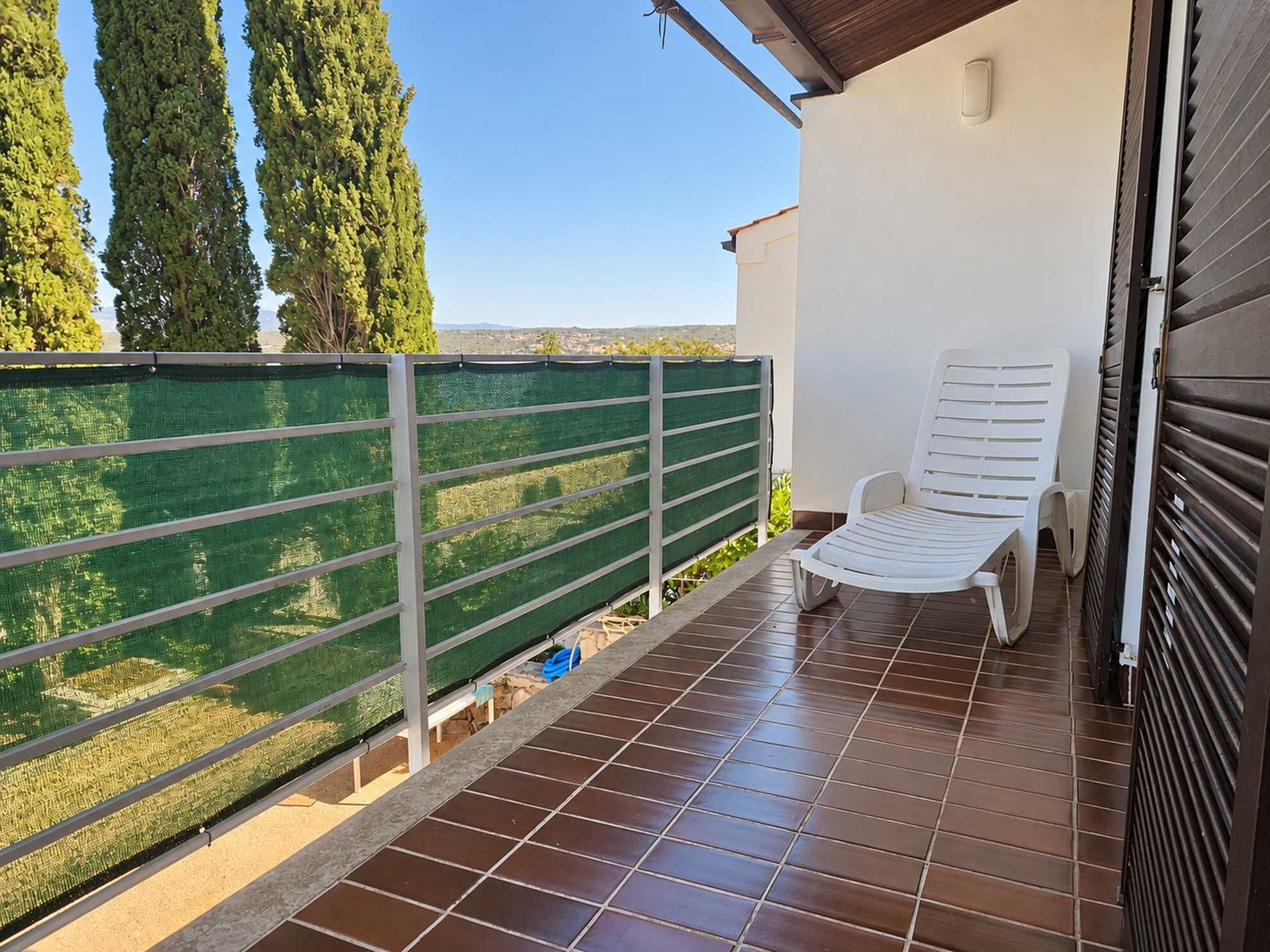Varanda com cadeira branca, cerca de rede verde e vista para árvores e paisagem distante.