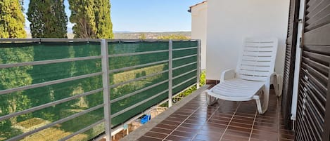 Un balcón con una silla blanca, una cerca de malla verde y vistas a árboles y un paisaje lejano.