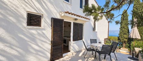 A white building with a patio area featuring a table and chairs, a view of the sea, and a pergola with a lamp.