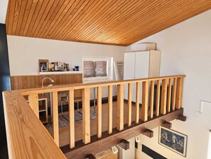 A loft-style room with a wooden ceiling, a balcony with a railing, a kitchen area with a refrigerator, and a window with blinds. - Apartment Hlebec with a Seaview (Vantačići)