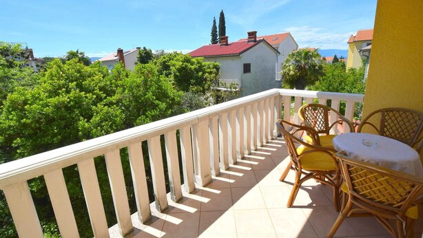 Un balcon doté d’une balustrade blanche, de deux fauteuils en osier, d’une petite table ronde et offrant une vue sur un quartier résidentiel avec des maisons aux toits rouges et des arbres verdoyants.