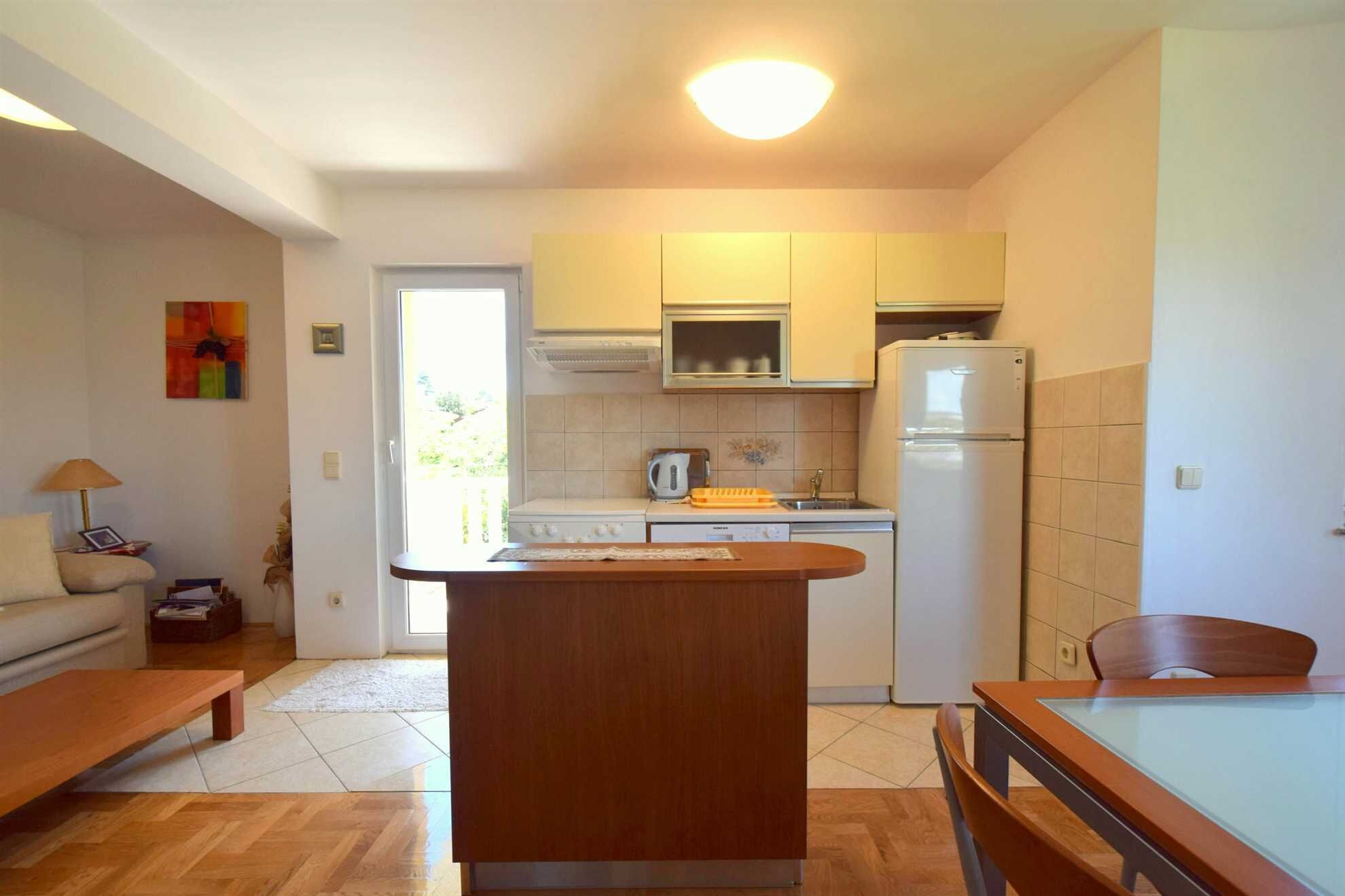 Een compacte keuken met een centraal eiland, een eettafel en een bank in een lichte kamer met een deur naar buiten.