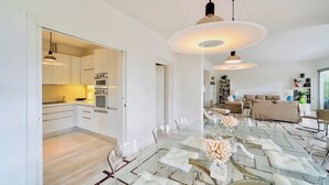 Une cuisine moderne avec des meubles blancs et des appareils en acier inoxydable, un coin repas avec une table et des chaises en verre, et un salon avec un canapé et des étagères.