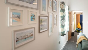 A hallway with framed artworks on the walls, a door leading to another room, and a couch in the foreground. - Ty Cwyfan: 3‑Bed Ground-Floor Apartment in Aberffraw | Managed by Anglesey Holiday Lettings (Ty Croes)