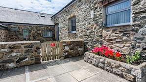 A stone building with a gated entrance, stone walls, and a flower bed with red flowers. - Ty Cwyfan: 3‑Bed Ground-Floor Apartment in Aberffraw | Managed by Anglesey Holiday Lettings (Ty Croes)