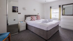 A modern hotel room with a large bed, bedside tables, a mirror, and a window with curtains. - Ty Cwyfan: 3‑Bed Ground-Floor Apartment in Aberffraw | Managed by Anglesey Holiday Lettings (Ty Croes)