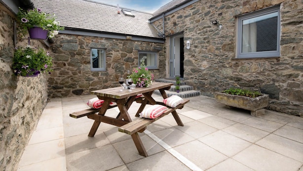 A stone building with a tiled patio, a wooden table with cushions, and potted plants. - Ty Cwyfan: 3‑Bed Ground-Floor Apartment in Aberffraw | Managed by Anglesey Holiday Lettings (Ty Croes)