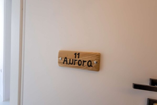 Een houten bord met het nummer 11 en de tekst 'Aurora' op een witte muur.