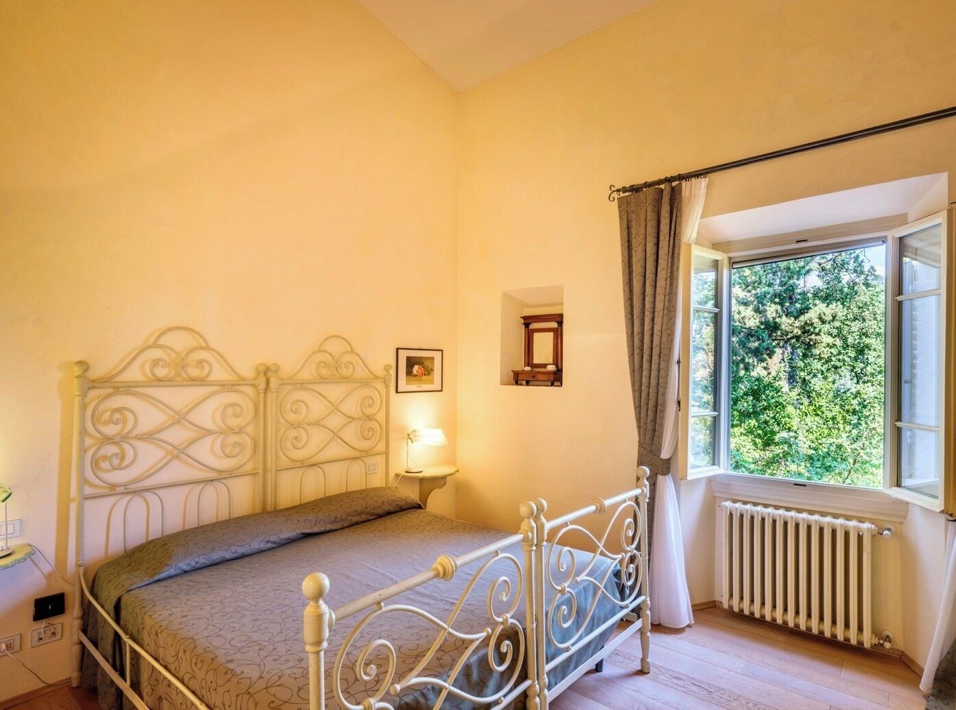 A bedroom with a metal bed frame, a bedside table with a lamp, a window with curtains, and a radiator.
