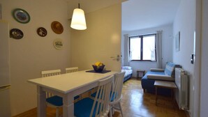 Un comedor con una mesa blanca y sillas azules, un frutero sobre la mesa y una ventana con cortinas.