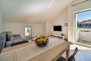 En moderne stue med sofa, spisebord med skål med frugt, fladskærms-tv og udsigt til det fri via en glasdør.