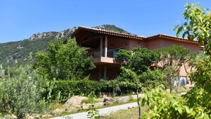 A two-story building with a balcony, surrounded by greenery and a mountainous backdrop. - Olmeto Plage: Large 3-bedroom apartment 200 metres from the sea (Olmeto)