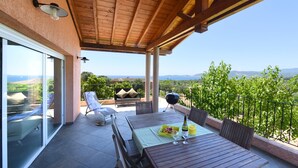 A covered patio with a wooden table set for dining, a glass door leading to a pool area, and a view of the landscape. - Olmeto Plage: Large 3-bedroom apartment 200 metres from the sea (Olmeto)