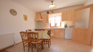 Een keuken met houten kastjes, een eettafel met stoelen, een klok aan de muur en een raam met gordijnen.