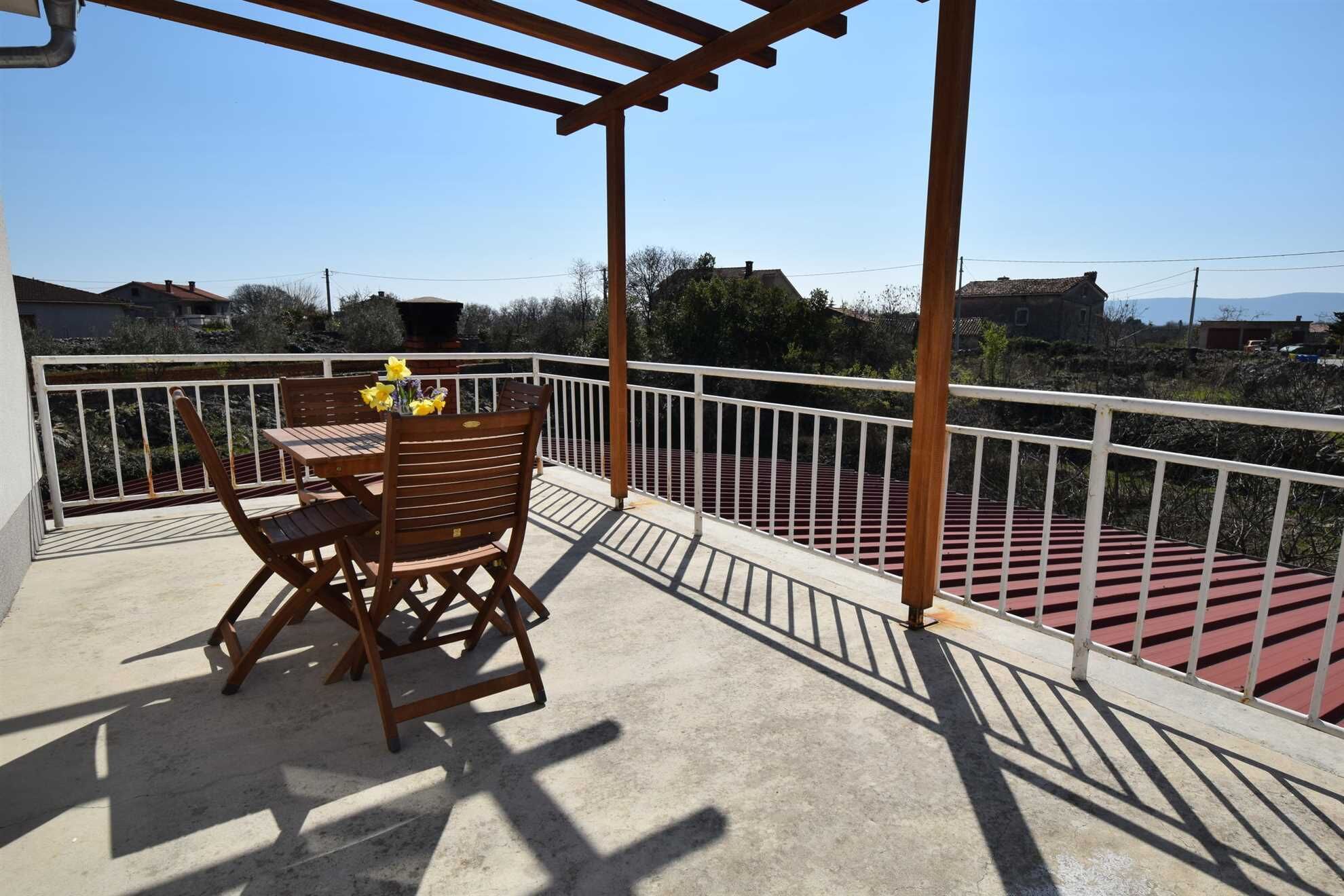 A balcony with a wooden table and chairs, a white railing, and a view of the surrounding landscape.