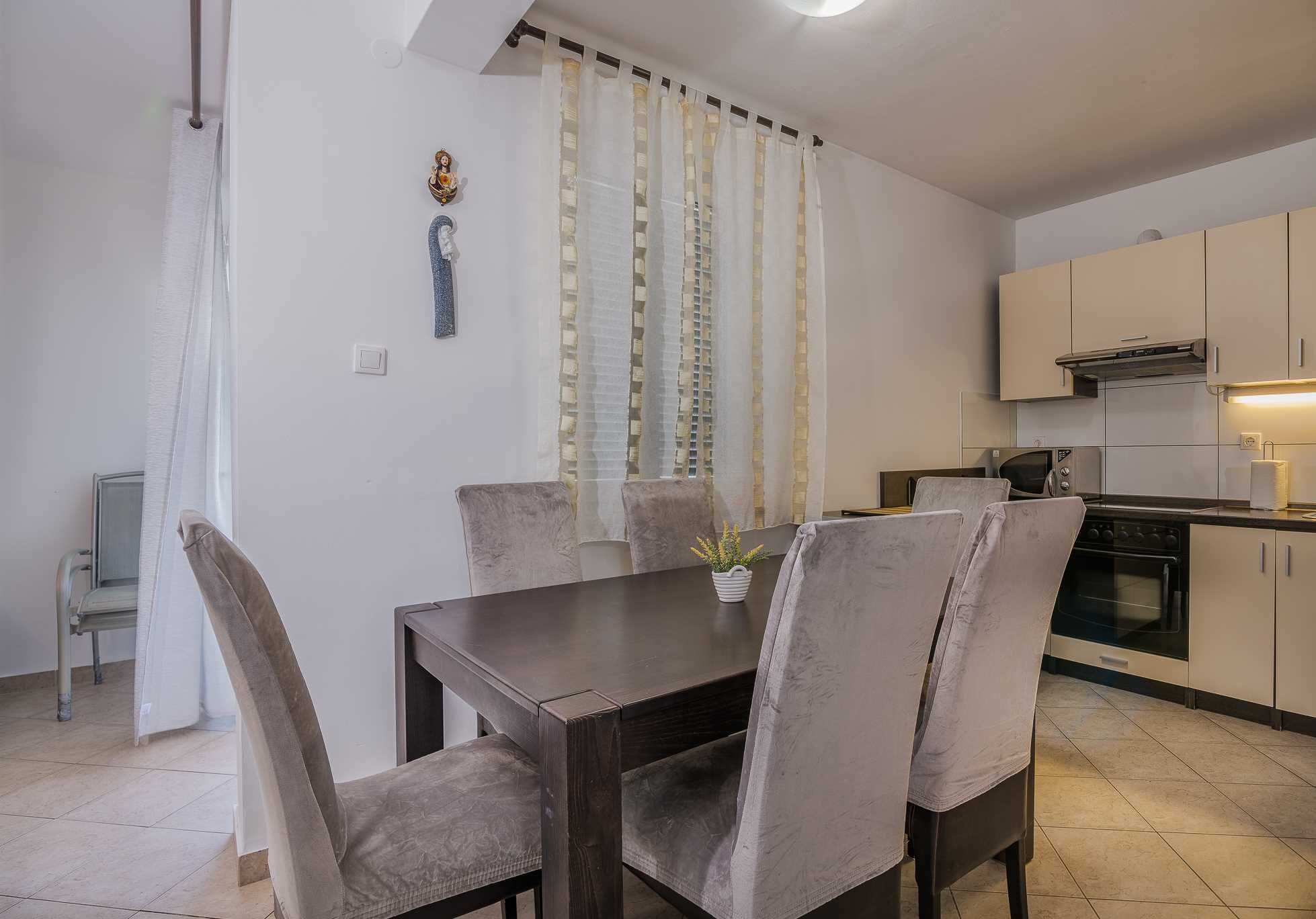 A dining area with a dark wooden table and cushioned chairs, a kitchen with white cabinets and appliances, and a window with sheer curtains.