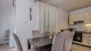 A dining area with a dark wooden table and cushioned chairs, a kitchen with white cabinets and appliances, and a window with sheer curtains. - Apartment Šop with balcony (Čižići)