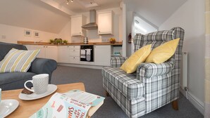 A cozy living room with a grey sofa, a plaid armchair with yellow cushions, a coffee table with a book and a mug, and a kitchen in the background. - Ty Mabon: 1‑Bed First-Floor Apartment in Aberffraw | Managed by Anglesey Holiday Lettings (Ty Croes)