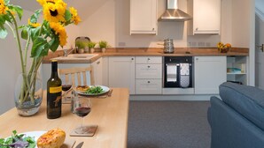 A modern kitchen with white cabinets, a stainless steel range hood, and a wooden countertop. There is a dining table with a bottle of wine, a glass of wine, and a plate of food. - Ty Mabon: 1‑Bed First-Floor Apartment in Aberffraw | Managed by Anglesey Holiday Lettings (Ty Croes)