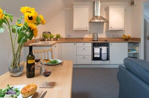 A modern kitchen with white cabinets, a wooden countertop, and a dining area with a table set for a meal. - Ty Mabon: Pet-friendly, first floor apartment in Aberffraw (Ty Croes)