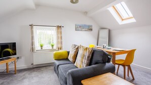 A modern living room with a grey sofa, yellow and grey cushions, a wooden coffee table, a dining table with a yellow chair, a TV on a wooden stand, a window with curtains, a radiator, and a skylight. - Ty Mabon: 1‑Bed First-Floor Apartment in Aberffraw | Managed by Anglesey Holiday Lettings (Ty Croes)