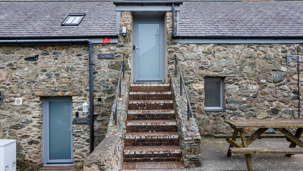 A stone building with a grey door, a staircase leading to a loft, and a picnic table outside. - Ty Mabon: 1‑Bed First-Floor Apartment in Aberffraw | Managed by Anglesey Holiday Lettings (Ty Croes)