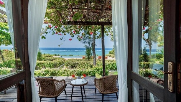 Une terrasse en bois, agrémentée de chaises en osier et d’une petite table, qui offre une vue imprenable sur un jardin luxuriant et l’océan.