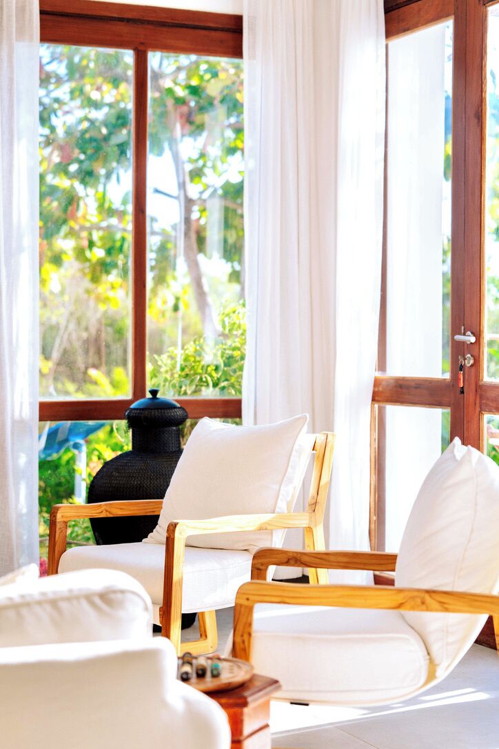 A room with a wooden-framed window, white curtains, and a view of greenery outside. There are two white chairs with cushions.