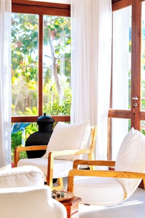 Habitación con ventana de madera, cortinas blancas y vista a zonas verdes. Se observan dos sillones blancos con cojines.