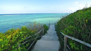 Un chemin de béton mène à une plage où l’eau est d’un bleu turquoise éclatant, et une clôture en bois borde le chemin d’un côté.