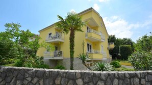 A two-story yellow building with balconies, surrounded by greenery and a stone wall.