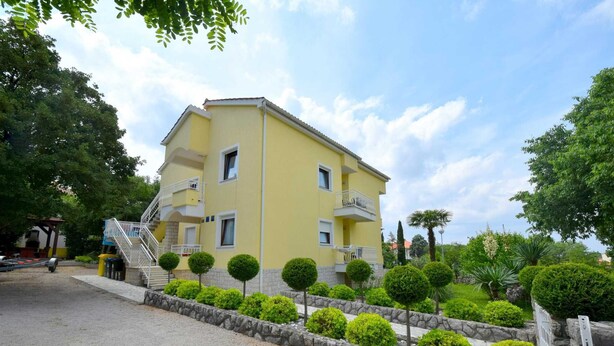 A two-story yellow building with a stone retaining wall and manicured bushes.