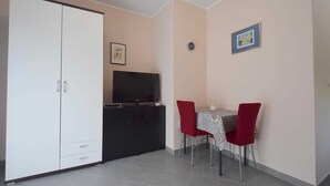 A small room with a white wardrobe, a TV on a black stand, a dining table with two red chairs, and a small white table.