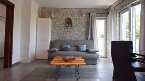 Un salon avec un mur en pierre, un canapé gris agrémenté de coussins, une table basse en bois et un fauteuil noir.