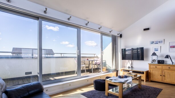 A modern living room with a large sliding glass door leading to a balcony, a wooden dining table, a black leather sofa, and a TV mounted on the wall. - Minstrel Lodge : Spacious 4‑Bed Pet-Friendly Villa with Sea Views in Rhosneigr | Managed by Anglesey (Rhosneigr)