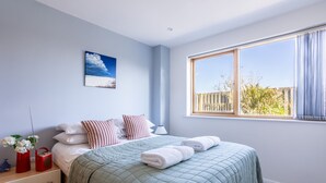 A bedroom with a large window, a bed with a green quilt, white pillows, and a red vase with flowers. - Minstrel Lodge : Spacious 4‑Bed Pet-Friendly Villa with Sea Views in Rhosneigr | Managed by Anglesey (Rhosneigr)