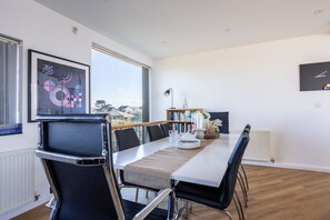 A modern dining area with a long table, black chairs, a bookshelf, and a large window offering a view.
