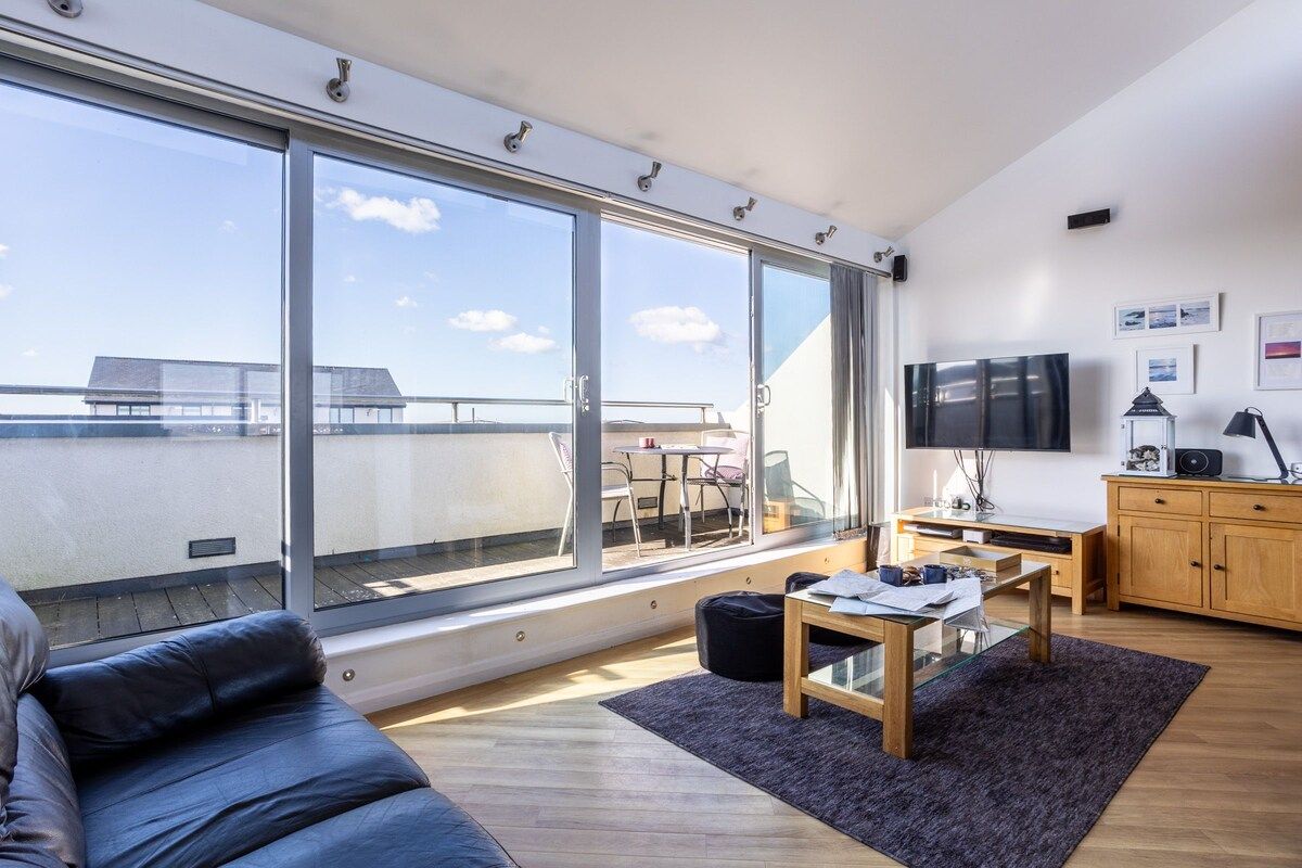 A modern living room with a large sliding glass door leading to a balcony, a flat-screen TV, a wooden cabinet, and a comfortable seating area.