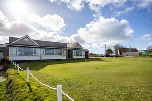 A building with a large windowed facade, a grassy area in front, and a clear sky. - Minstrel Lodge : Spacious 4‑Bed Villa Rhosneigr (Rhosneigr)