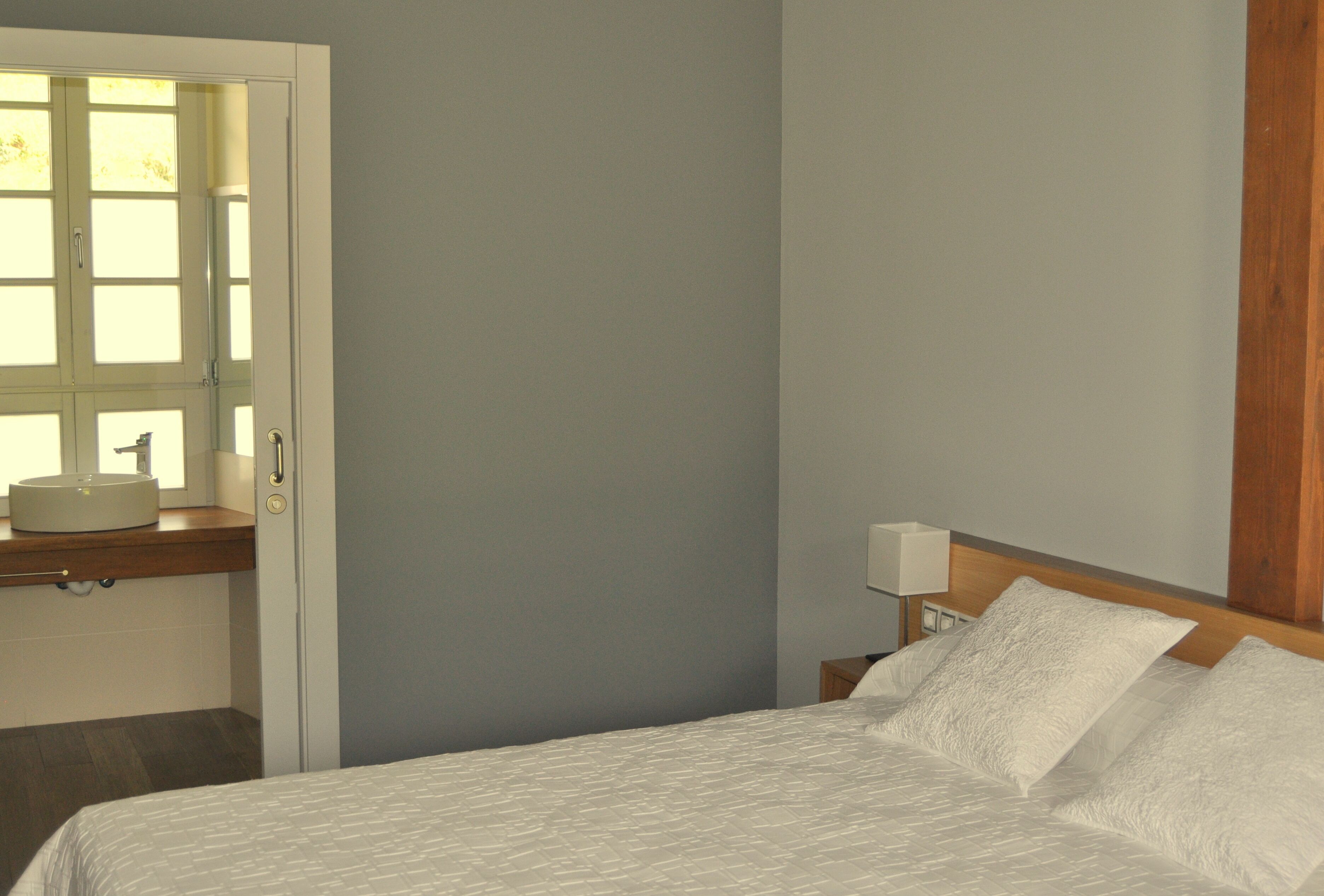 A hotel room with a bed, a bedside table with a lamp, a window with a sink, and a wooden headboard.