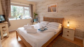 A bedroom with a wooden bed, white linens, a window with curtains, a painting on the wall, and a bedside table with a lamp.