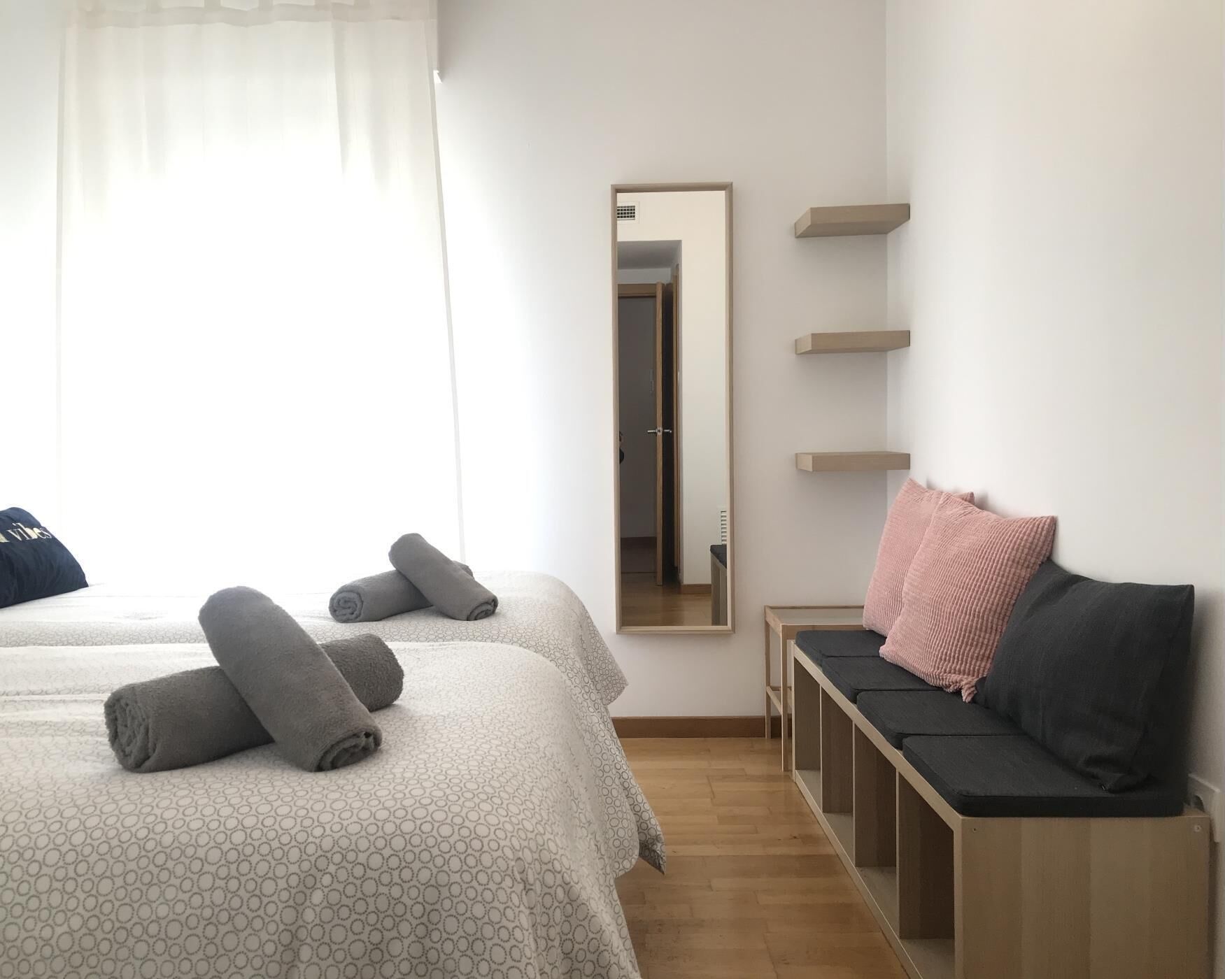 Un dormitorio con dos camas, un banco, un espejo y una ventana con cortinas.