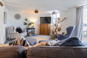 A living room with a sofa, armchair, TV, wooden cabinet, and decorative plants. - Driftwood: Pet-friendly seaside home for six in Rhosneigr (Rhosneigr)