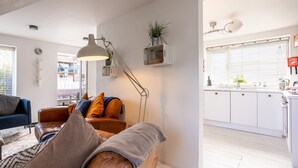 A modern living room with a brown leather sofa, a blue armchair, and a floor lamp. A kitchen with white cabinets and a window with blinds is visible in the background. - Driftwood : Pet‑Friendly Seaside Home in Rhosneigr | Managed by Anglesey Holiday Lettings (Rhosneigr)