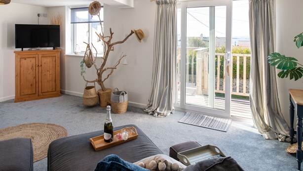 A living room with a sofa, a wooden cabinet, a TV, a plant, and a balcony with curtains. - Driftwood : Pet‑Friendly Seaside Home in Rhosneigr | Managed by Anglesey Holiday Lettings (Rhosneigr)