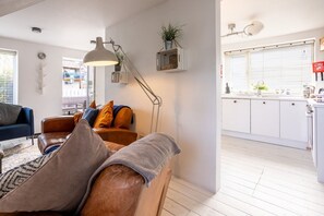 A modern living room with a sofa, a floor lamp, and a kitchen area with white cabinets and a window. - Driftwood: Pet-friendly seaside home for six in Rhosneigr (Rhosneigr)