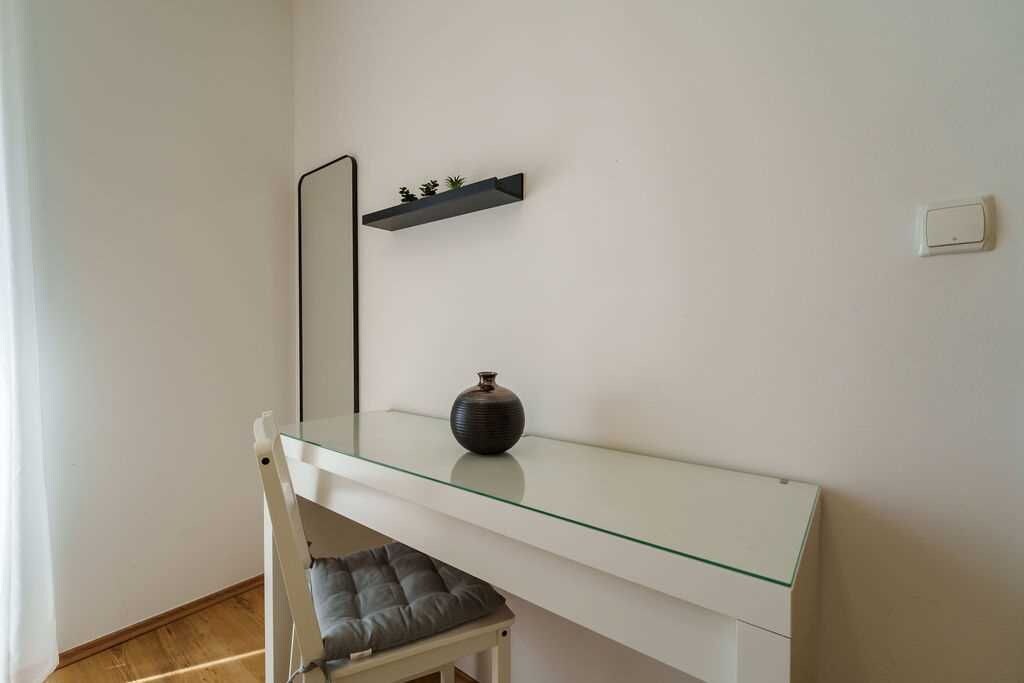Una habitación con un escritorio de superficie de vidrio, una silla, un estante pequeño con plantas, un florero y un interruptor de luz en la pared.