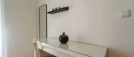 A room with a glass-top desk, a chair, a small shelf with plants, a vase, and a wall-mounted light switch.