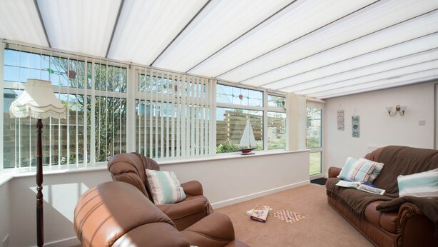 Ein sonnendurchflutetes Zimmer mit zwei Ledersofas, einer Stehlampe und Blick auf einen Garten durch große Fenster.